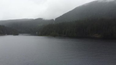 Calm lake surrounded by green forest hills with a fog on a rainy day