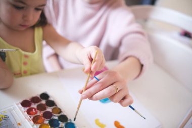 Anne ve kızı evdeki beyaz bir masada suluboya fırçalarla boya yapıyorlar.