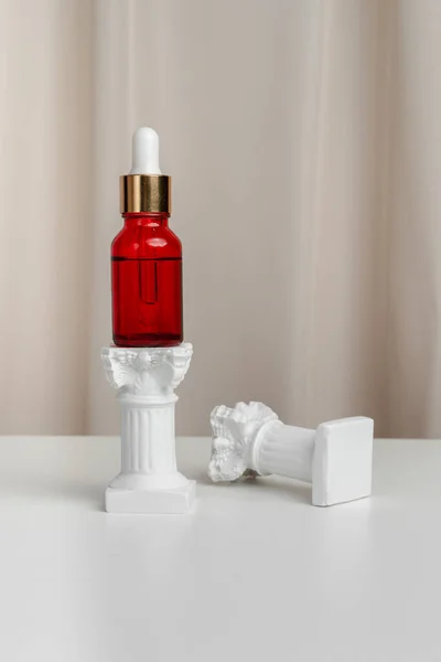 Face serum in a red bottle stands on a pillar. Classic style. Mockup for advertising your product. Essence for skin health. Anti-wrinkle serum on a beige fabric with beautiful folds.
