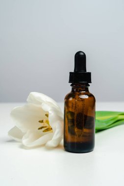 Koyu cam bir kavanozda masaj yağı. Cilt sağlığı için esans. Moda tarzı. Ürününün modeli. Kavramsal fotoğraf. Beyaz arka planda Aroma yağı ve laleler.