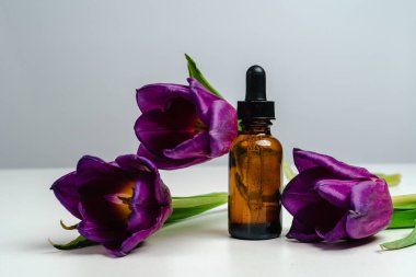 Koyu cam bir kavanozda masaj yağı. Cilt sağlığı için esans. Moda tarzı. Ürününün modeli. Kavramsal fotoğraf. Beyaz arka planda Aroma yağı ve laleler. Erkekler ve Kadınlar için Unisex Kozmetikleri