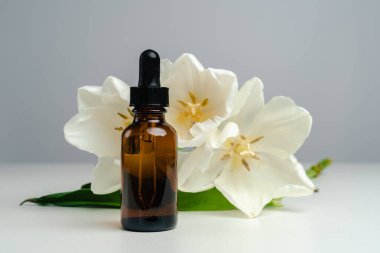 Koyu cam bir kavanozda masaj yağı. Cilt sağlığı için esans. Moda tarzı. Ürününün modeli. Kavramsal fotoğraf. Beyaz arka planda Aroma yağı ve laleler.