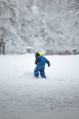 Mavi kışlık ceketli mutlu çocuk ağaçlar arasında karda oynamaktan hoşlanıyor. Kar taneleri düşüyor..