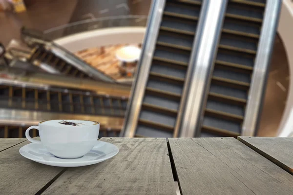 Defocus with terrace wood and cappuccino coffee with escalators in shopping mall for background usage — Stock Photo, Image