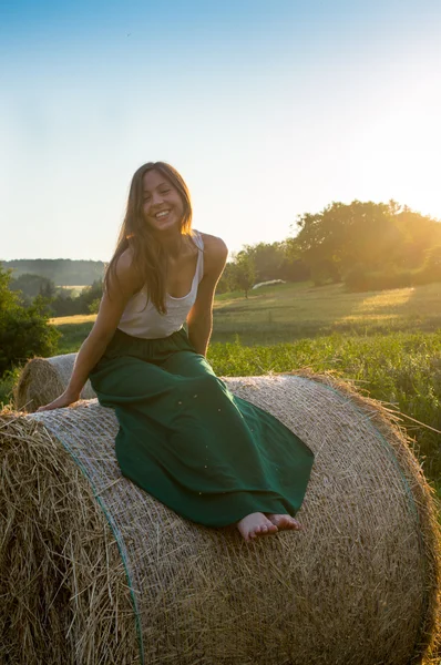 Hermosa chica sentada en la paca redonda de heno en la puesta del sol — Foto de Stock