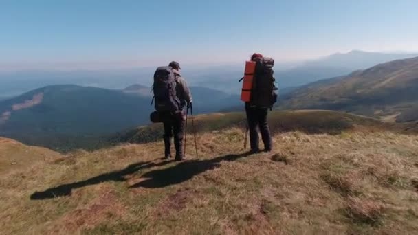 Man en vrouw met rugzakken en wandelstokken aan de rand van de klif kijken naar het bergmeer en de bergen aan de horizon — Stockvideo