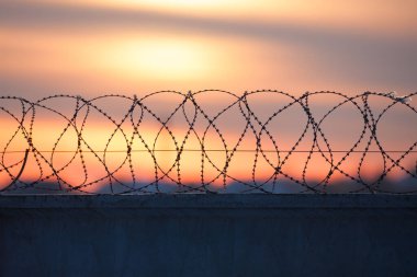 Razor wire fence silhouette against a dramatic sunrise or sunset sky. A powerful security and border concept symbolizing danger, prison, and restriction.
