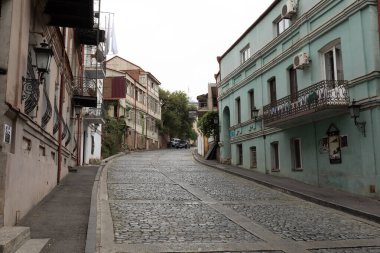Eski Tiflis, Gürcistan 'daki geniş bir kaldırım taşı caddesinin manzarası, bulutlu bir günde öne çıkan bir nane yeşili cephe de dahil olmak üzere yenilenmiş tarihi binalar tarafından çerçevelenmiş..