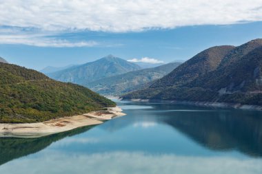Mavi gökyüzünü yansıtan ve Gürcistan 'da sık ormanlarla çevrili dağlarla çevrili turkuaz dağ rezervuarının çarpıcı panoramik görüntüsü..