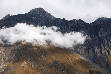Koyu kayalık dağ zirvelerinde beyaz bulutlar ve sis bulunur. Engebeli dik yamaçlar kasvetli bir yabanda kahverengi sonbahar renklerini gösterir.