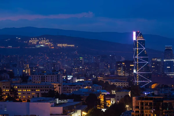 Gürcistan 'ın Tiflis kentinin geniş gece manzarası alacakaranlıkta parlayan geniş bir şehre karşı geometrik aydınlatma ile yükselen modern bir gökdeleni vurguluyor..