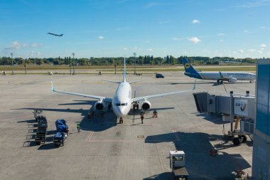 Boryspil, Ukraine - September 30, 2018: Commercial aircraft is being serviced by ground crew and utility vehicles on a wide airport tarmac, with another airplane taking off in the background on a sunny day.