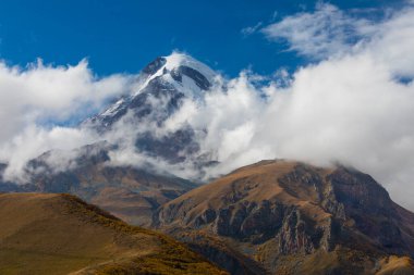 Gürcistan 'ın Kafkasya Dağları' ndaki beyaz bulutların ve engebeli sonbahar tepelerinin üzerinde yükselen Kazbek Dağı 'nın çarpıcı manzarası..