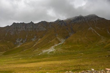 Engebeli, puslu tepeler ve yeşil ve kahverengi sonbahar otlarıyla kaplı geniş, dokulu yamaçlar içeren dramatik bir Kafkasya dağ manzarası..
