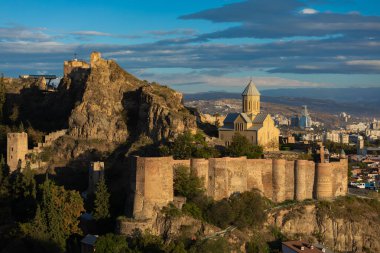 Gürcistan 'ın Tiflis kentindeki antik Narikala Kalesi ve St. Nicholas Kilisesi' nin manzarası, mavi bulutlu gökyüzüne karşı sıcak altın güneş ışığıyla aydınlanıyor..