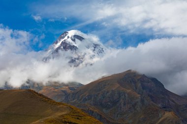 Gürcistan 'ın Kafkasya Dağları' ndaki mavi gökyüzüne karşı Kazbek Dağı 'nın karlı zirvesi bulutların üzerinde yükseliyor ve sonbahar renkli tepeler üzerinde yükseliyor..