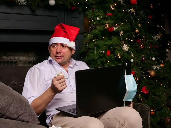 A sick man alone greets the new year. A man with a laptop next to a Christmas tree.