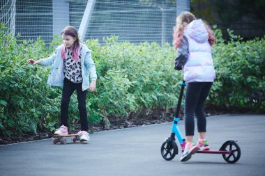 Aktif kızlar kaykay parkında scooter ve kaykay kullanıyor. Çocuklar boş vakitlerinde oynuyorlar. Sağlıklı yaşam tarzı kavramı. Çocuk sporu.
