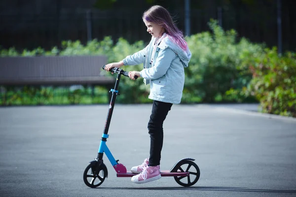 Szczęśliwa piękna dziewczyna jedzie ulicą na hulajnodze w wietrzny dzień. Dziecko bawi się wolnym czasem. Koncepcja zdrowego stylu życia. Sport dla dzieci — Zdjęcie stockowe