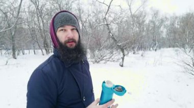 Sakallı bir adam kış yürüyüşünde kahve içer, bir kupadan kar üfler, kar yağar.