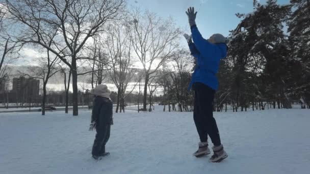 Mãe e filha, alegremente jogando neve uns nos outros enquanto estavam em uma estrada coberta de neve através da floresta no inverno dia ensolarado gelado. Movimento lento. — Vídeo de Stock