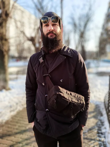 Porträt eines jungen bärtigen Europäers in Winterjacke und Hut, der in einer verschneiten Winterstadt spaziert — Stockfoto