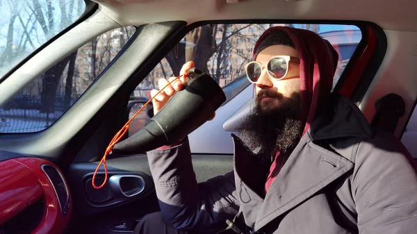 Retrato de cerca de un joven barbudo tomando café para llevar. Un barbudo con bigote mira por la ventana del coche. Hombre en el coche bebiendo café — Foto de Stock