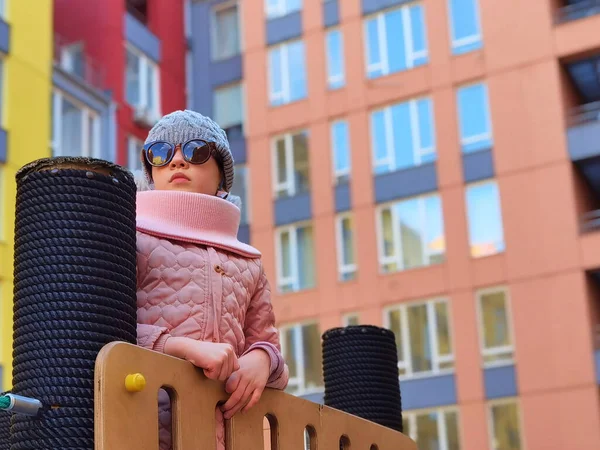 La niña de 9 años en el patio de recreo. El niño está vestido con ropa cálida de temporada, una chaqueta rosa y un sombrero gris.. — Foto de Stock