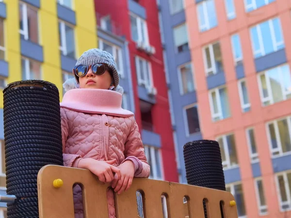 La niña de 9 años en el patio de recreo. El niño está vestido con ropa cálida de temporada, una chaqueta rosa y un sombrero gris.. — Foto de Stock