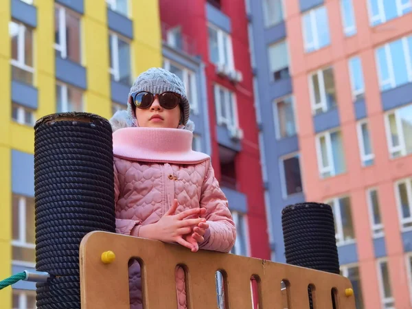 La niña de 9 años en el patio de recreo. El niño está vestido con ropa cálida de temporada, una chaqueta rosa y un sombrero gris.. — Foto de Stock