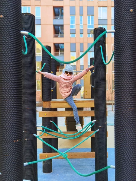 Das Mädchen 9 Jahre alt auf dem Spielplatz. Das Kind trägt warme Halbjahreskleidung, eine rosa Jacke und einen grauen Hut. — Stockfoto