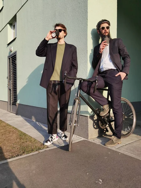 Dos jóvenes amigos vestidos casualmente pasar tiempo juntos en la ciudad, beber café para llevar próxima bicicleta. — Foto de Stock