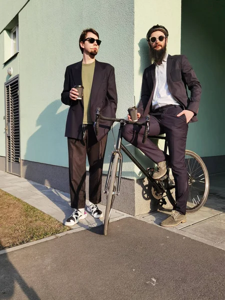 Dos jóvenes amigos vestidos casualmente pasar tiempo juntos en la ciudad, beber café para llevar próxima bicicleta. — Foto de Stock