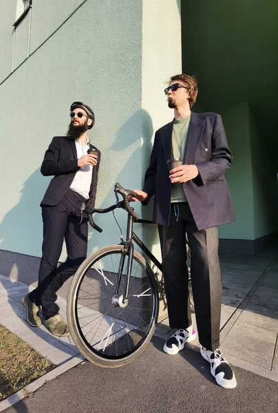 Two young men friends dressed casually spending time together at the city, drinking takeaway coffee next bike. — Stock Photo, Image