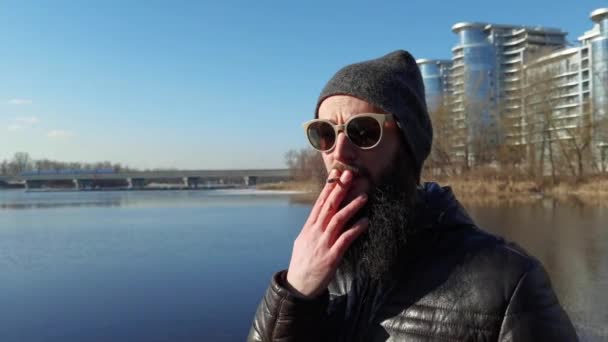 Un hipster fumando un cigarrillo al aire libre. Un joven fumando en la ciudad cerca de un edificio moderno o de un centro cultural. Malos hábitos y concepto de cultura juvenil. — Vídeos de Stock