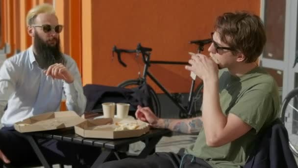 Charla amistosa. Dos jóvenes felices comiendo pizza y hablando entre sí mientras están sentados al aire libre — Vídeo de stock