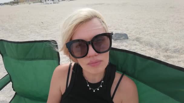 Slow motion portrait face of beautiful young woman with short blonde hair looking at camera sitting on sea beach — Stock Video