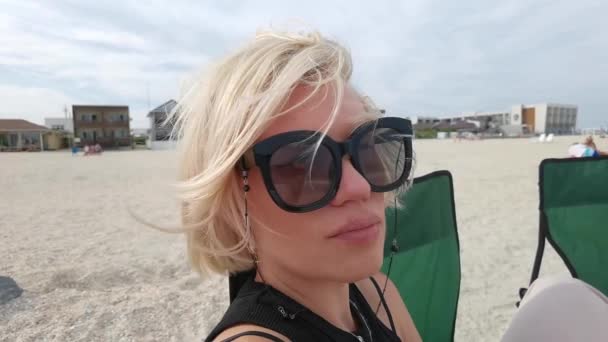 Портретное лицо красивой молодой женщины с короткими светлыми волосами, смотрящей на камеру, сидящую на морском пляже — стоковое видео