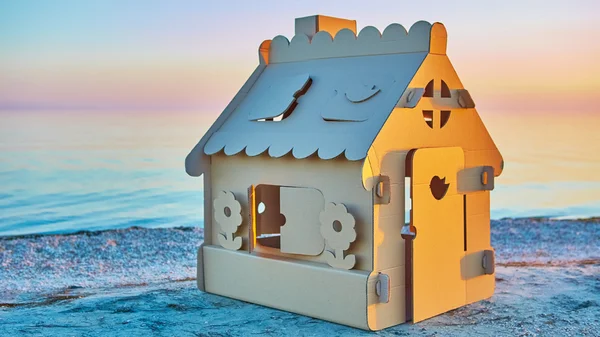 Casa de brinquedo feita de papelão ondulado na costa do mar ao pôr do sol . — Fotografia de Stock