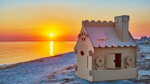 Casa de brinquedo feita de papelão ondulado na costa do mar ao pôr do sol . — Fotografia de Stock