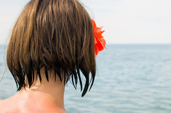 Menina com uma flor vermelha em seu cabelo castanho olhando para o mar — Fotografia de Stock