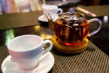 Cam çaydanlık, çiçek açan siyah çay, beyaz kupa masa ve koyu arka planda hayat..