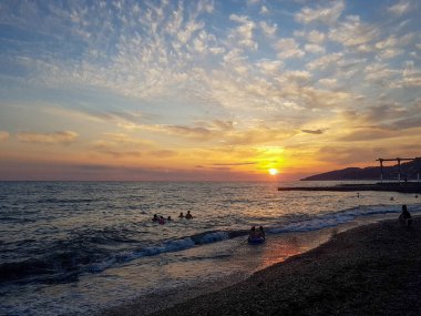 İnsanlar güneşleniyor, gün batımında kumlu tropikal kayalık plajda parti veriyorlar..