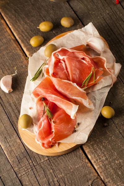 Closeup of thin slices of prosciutto with mixed olives and paprika on wooden cutting board — Stock Photo, Image