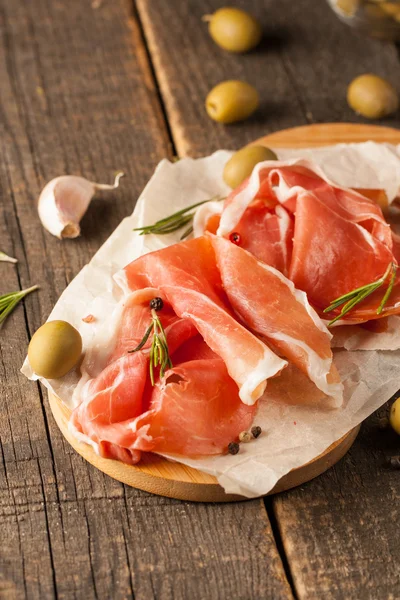 Closeup of thin slices of prosciutto with mixed olives and paprika on wooden cutting board — Stock Photo, Image