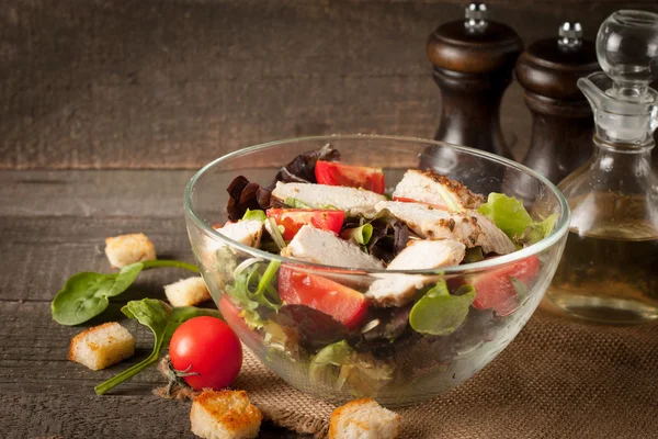 Salada fresca feita de tomate, rúcula, peito de frango, rúcula, biscoitos e especiarias. Salada Caesar em uma tigela branca e transparente em fundo de madeira — Fotografia de Stock