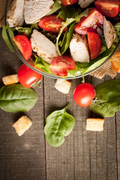 Salada fresca feita de tomate, rúcula, peito de frango, rúcula, biscoitos e especiarias. Salada Caesar em uma tigela branca e transparente em fundo de madeira — Fotografia de Stock