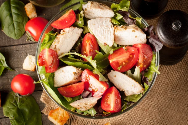 Fresh salad made of tomato, ruccola, chicken breast, arugula, crackers and spices. Caesar salad in a white, transparent bowl on wooden background — Stock Photo, Image