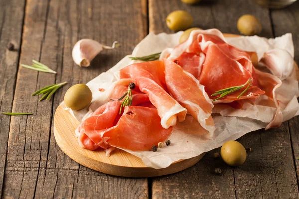 Closeup of thin slices of prosciutto with mixed olives and paprika on wooden cutting board