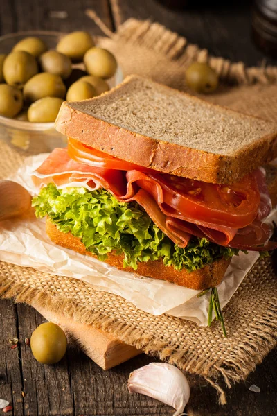 Close-up photo of a club sandwich. Sandwich with meet, prosciutto, salami, salad, vegetables, lettuce, tomato, onion and mustard on a fresh sliced rye bread on wooden background. Olives background.